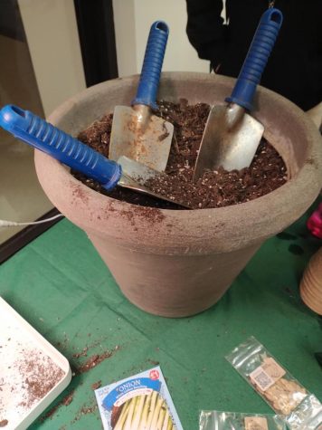 A terracotta pot rests upon the top of a green table cover with seeds strewn about it. The pot is filled with dirt. Three trowels with blue grips are wedged inside.